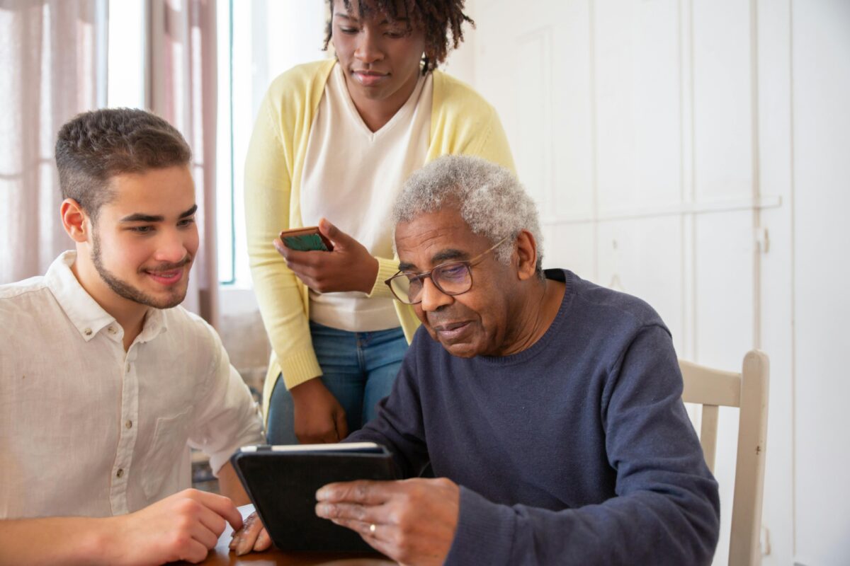 And older man on an ipad with a younger man next to him and a woman standing behind and looking.